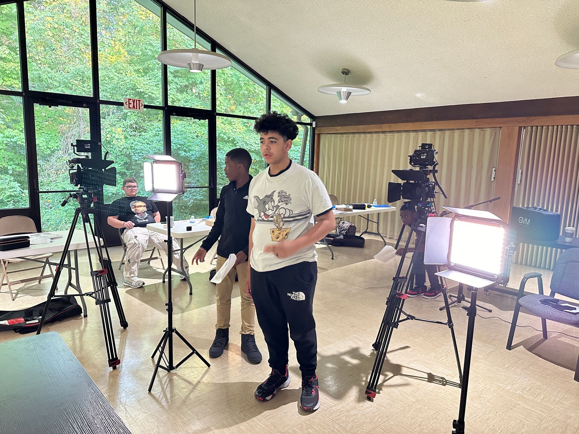 People setting up cameras and lighting equipment in a room with large windows and tables.