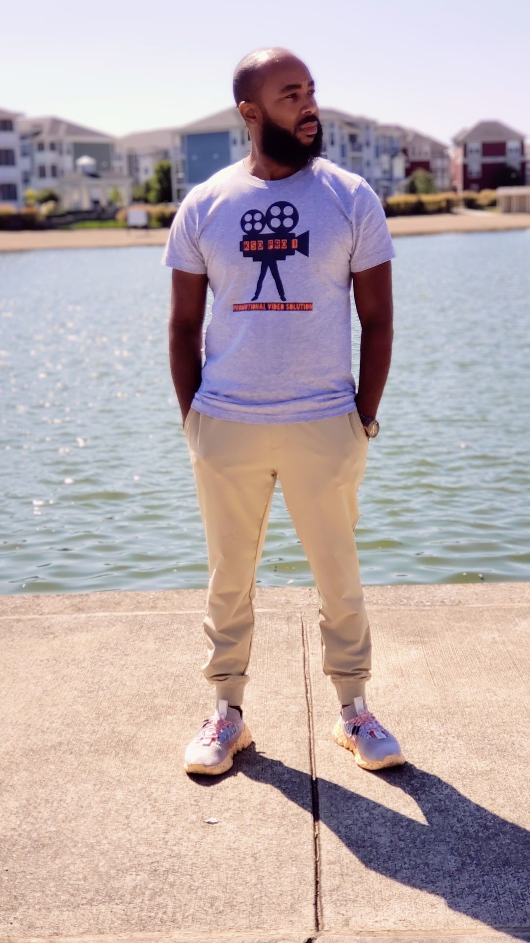 Man in a graphic t-shirt standing by a waterfront with buildings in the background.