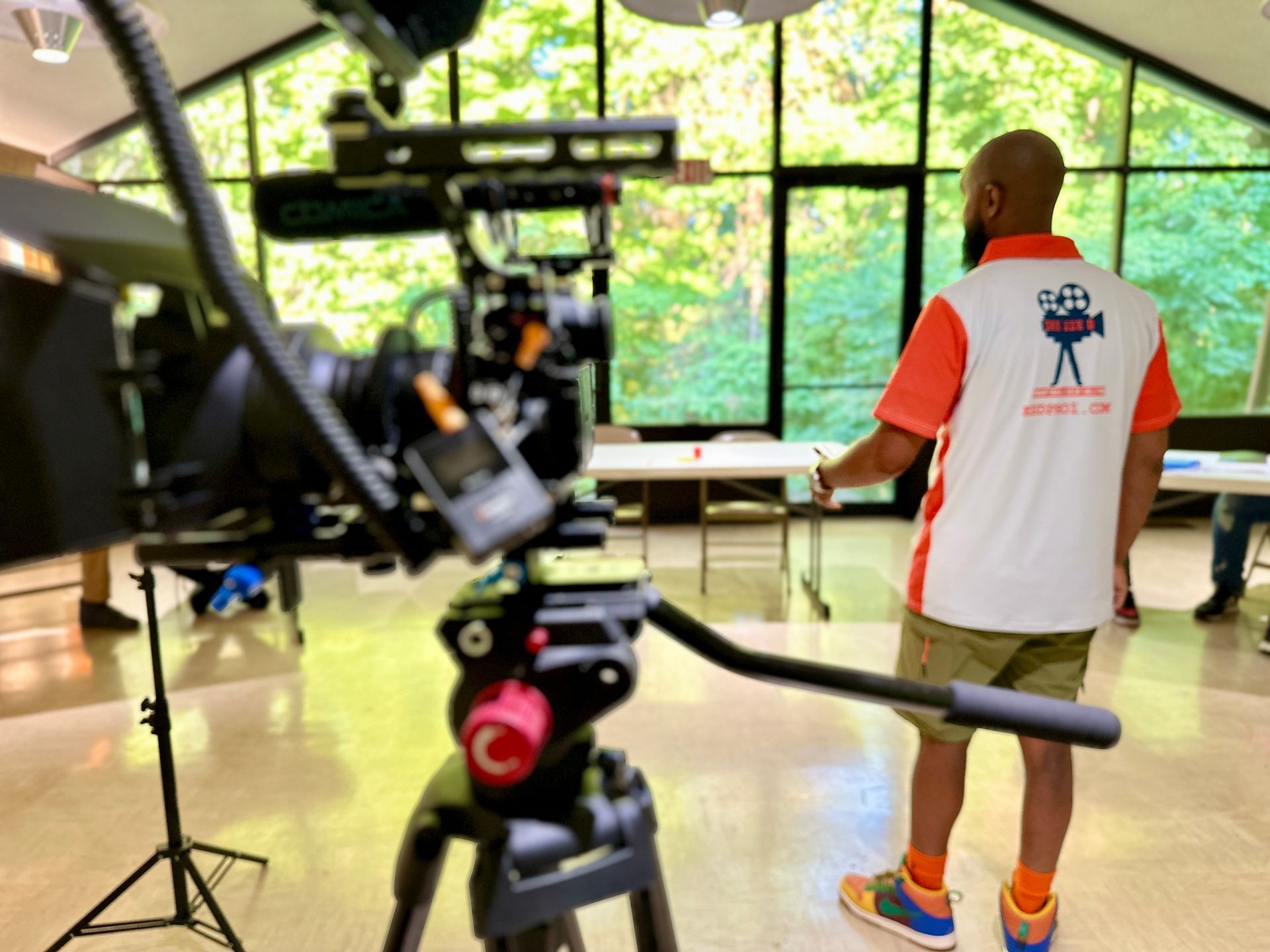 Man in sports attire standing near a large window, with a professional camera setup in the foreground.