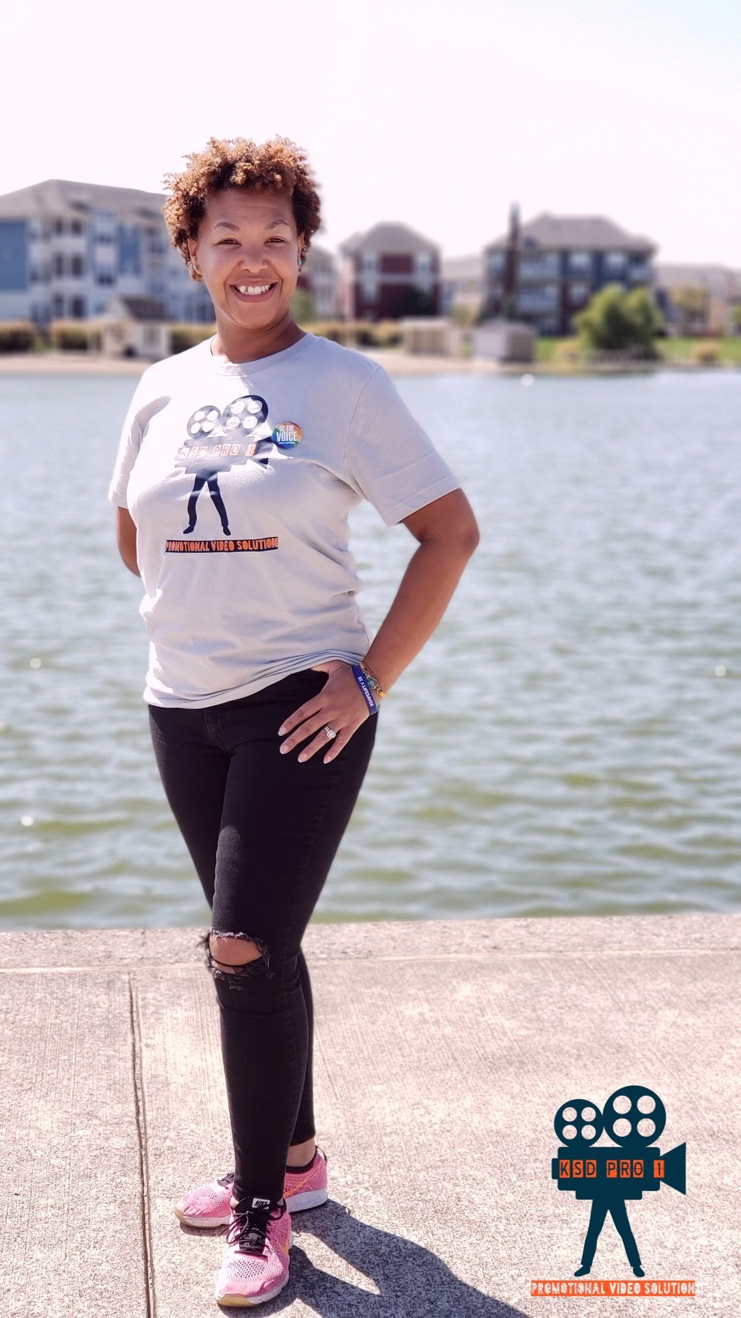 Smiling person standing by a lake, wearing a graphic t-shirt and pink sneakers.