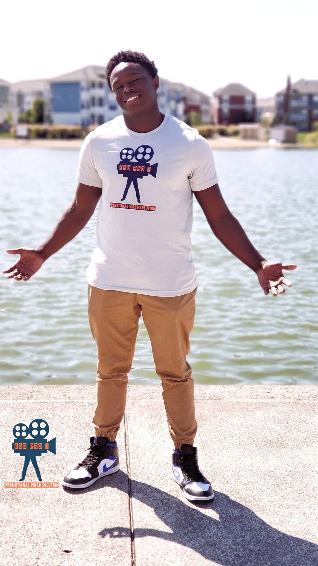 Person smiling in a logo t-shirt standing by a waterfront with buildings in the background.