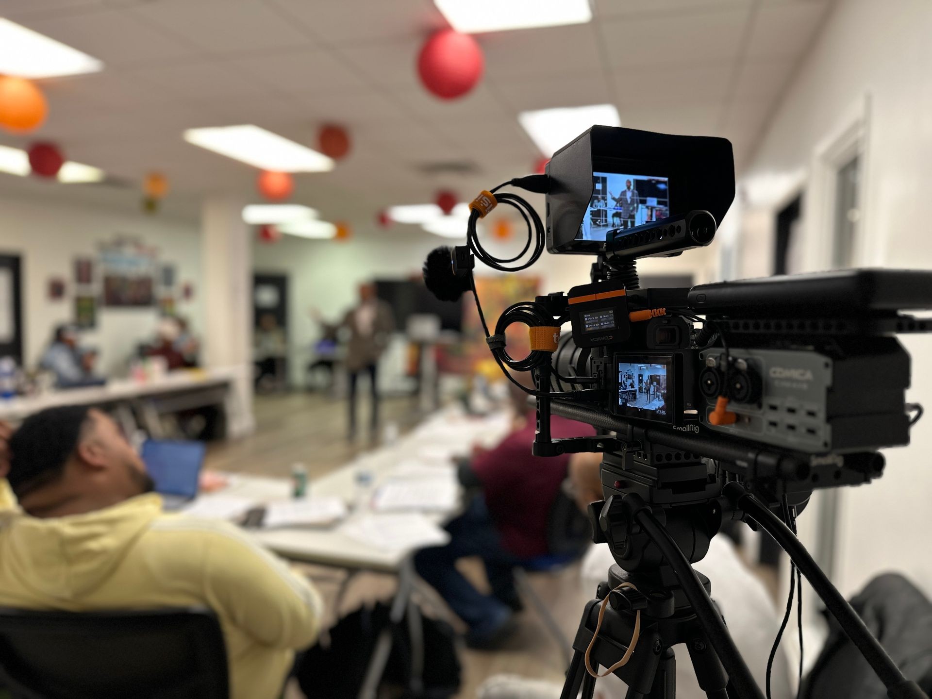 Video camera setup in a meeting room, with a blurred person speaking in the background.